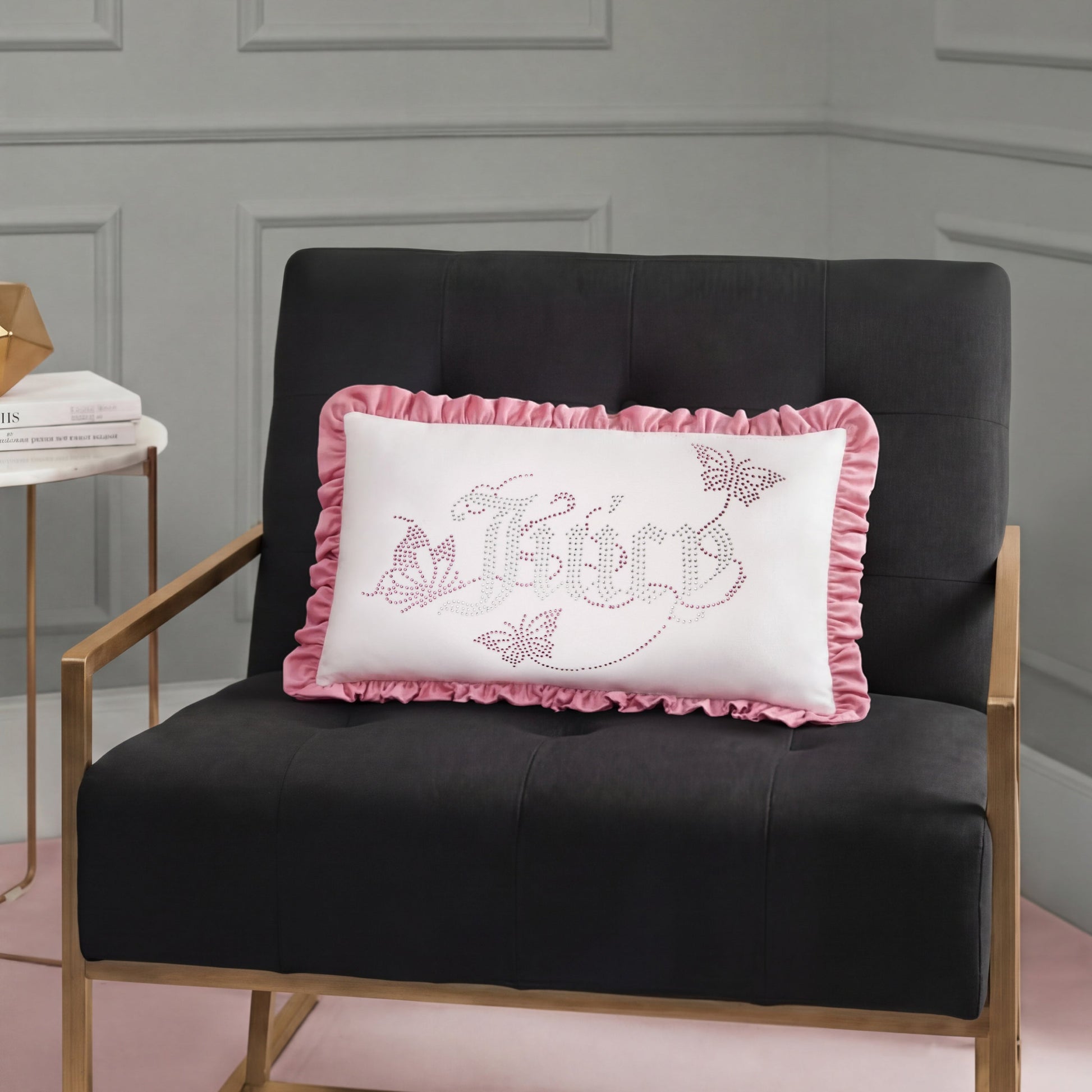 A black chair with gold armrests holds a white Ruffle Decorative Pillow featuring pink ruffles, "Believe" text, butterflies, and floral designs in a modern room with gray paneled walls.