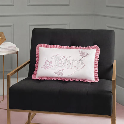 A black chair with gold armrests holds a white Ruffle Decorative Pillow featuring pink ruffles, "Believe" text, butterflies, and floral designs in a modern room with gray paneled walls.