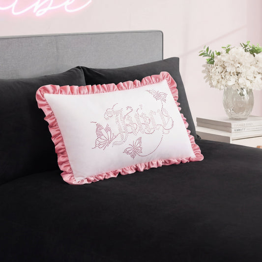 The Ruffle Decorative Pillow, featuring pink ruffled edges and cursive writing, rests on a black bedspread with a vase of white flowers displayed on the bedside table in the background.