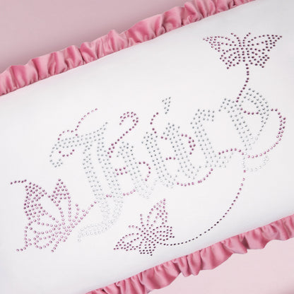 The Ruffle Decorative Pillow features “Jolie” and two butterflies in pink and purple rhinestones on a soft pink background with chic pink ruffled edges, perfect for adding a sparkly touch to your decor.