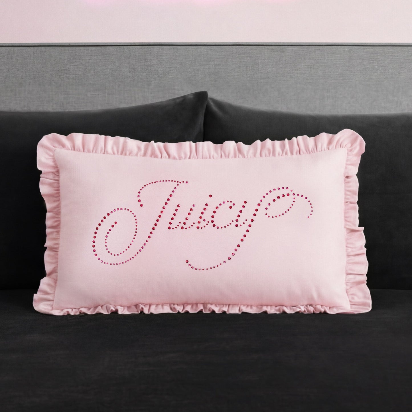 A rectangular pink Ruffle Decorative Pillow with ruffled edges sits on a dark couch. "Juicy" is written in cursive on the pillow with small, shiny pink studs for a glamorous touch.