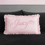 A rectangular pink Ruffle Decorative Pillow with ruffled edges sits on a dark couch. "Juicy" is written in cursive on the pillow with small, shiny pink studs for a glamorous touch.