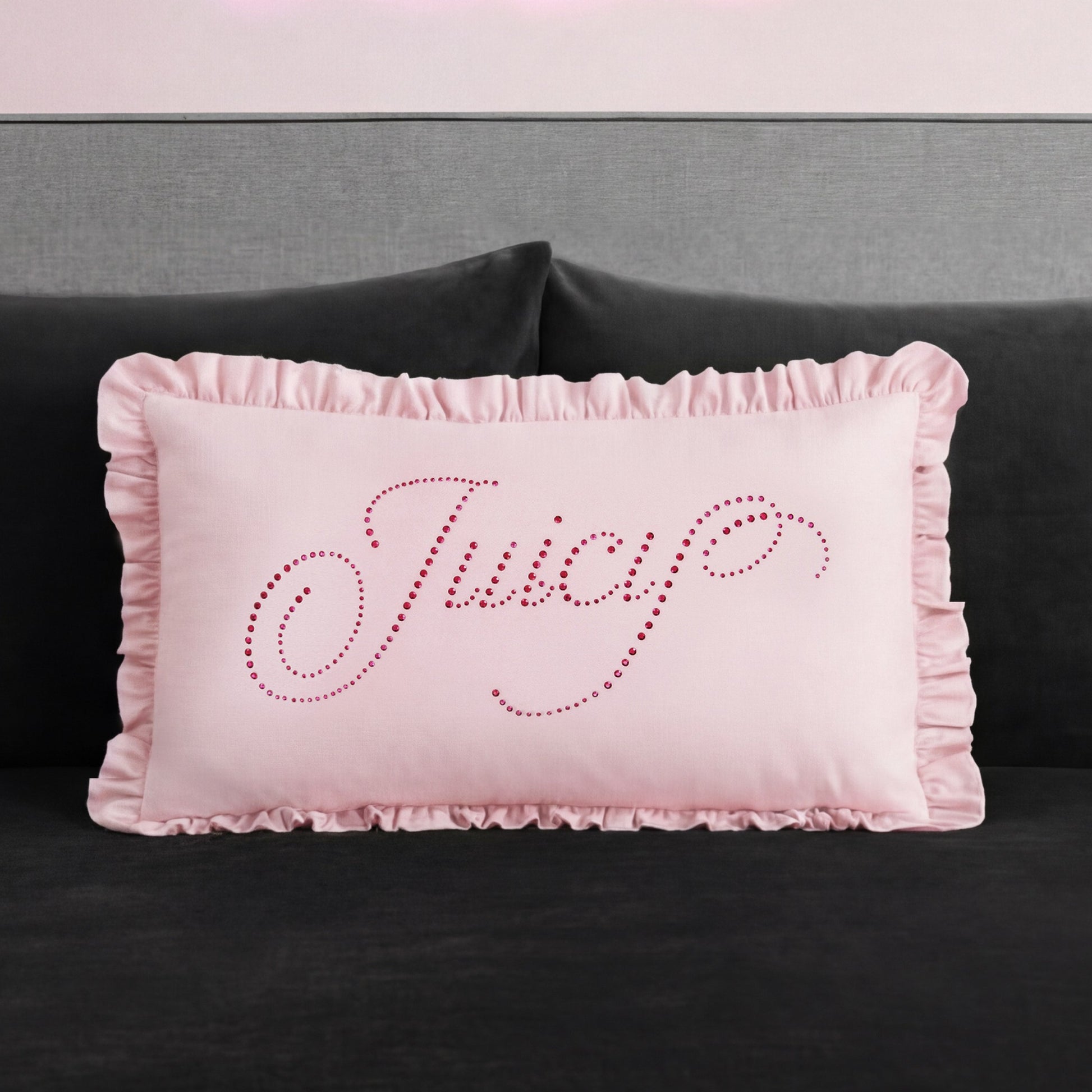 A rectangular pink Ruffle Decorative Pillow with ruffled edges sits on a dark couch. "Juicy" is written in cursive on the pillow with small, shiny pink studs for a glamorous touch.