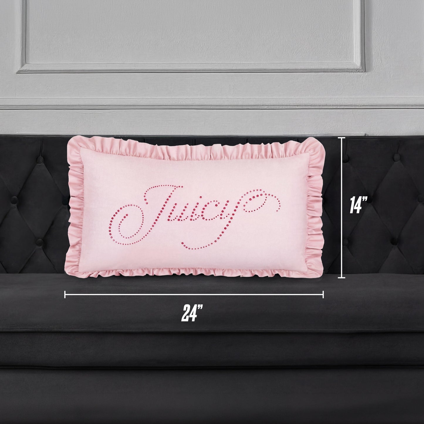 A Ruffle Decorative Pillow in pink with "Juicy" in cursive sparkles rests on a black tufted sofa. The pillow is 24"x14" and sits against a chic gray paneled wall for a stylish accent.
