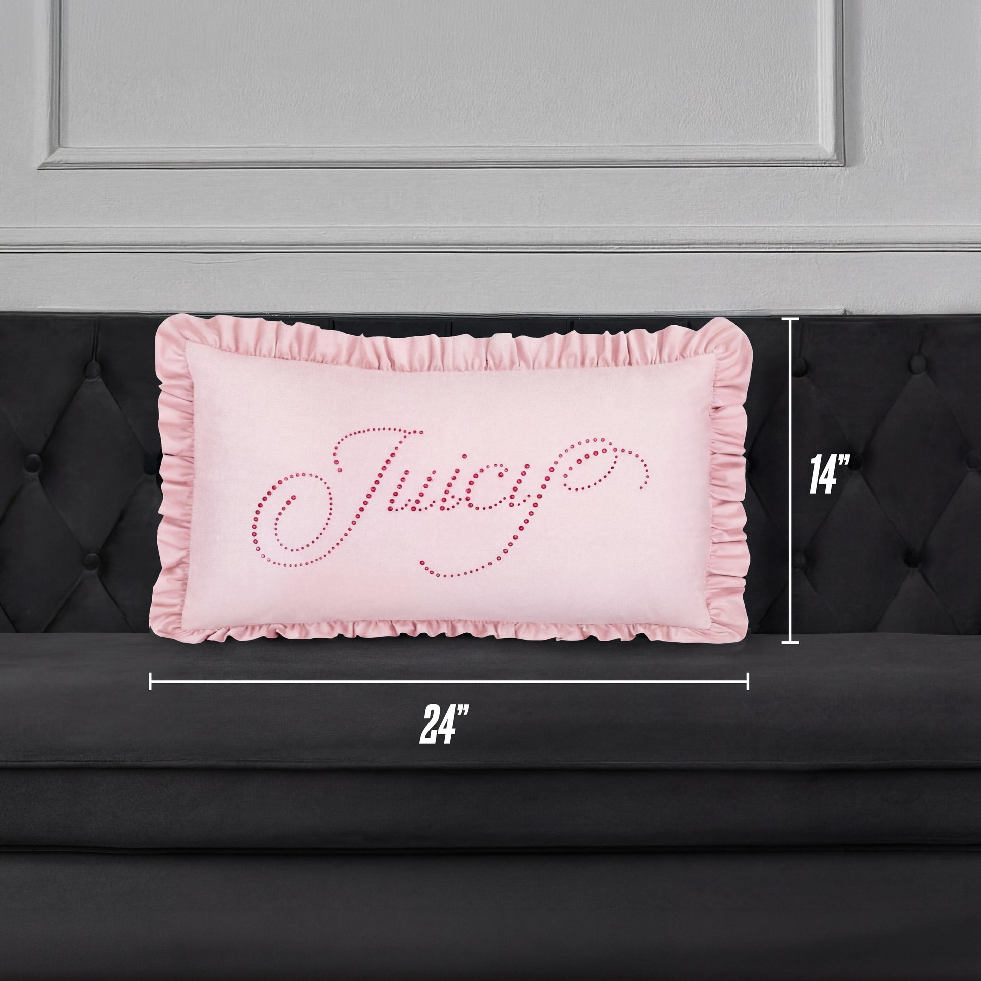 A Ruffle Decorative Pillow in pink with "Juicy" in cursive sparkles rests on a black tufted sofa. The pillow is 24"x14" and sits against a chic gray paneled wall for a stylish accent.
