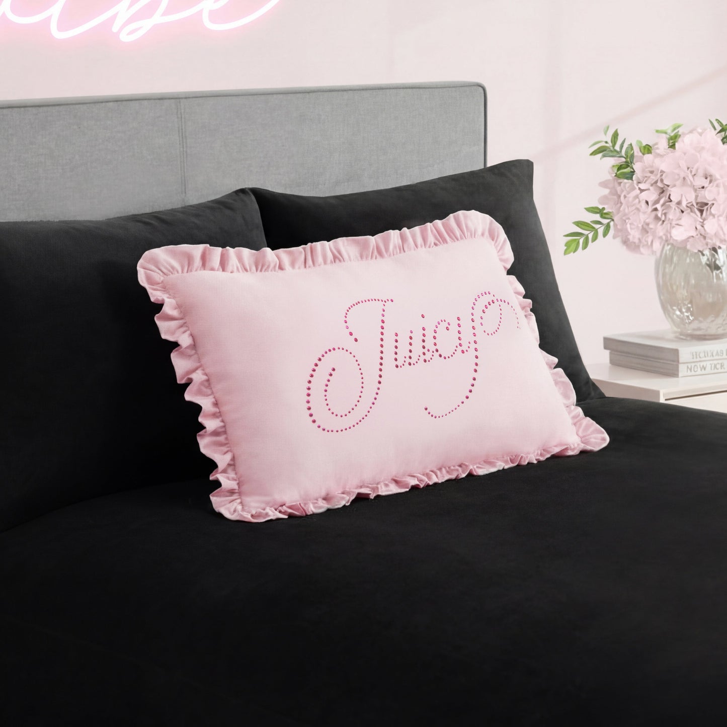 A pink Ruffle Decorative Pillow with rhinestone accents sits on a black bedspread, paired with a gray headboard and a vase of pink flowers on the bedside table in the background.