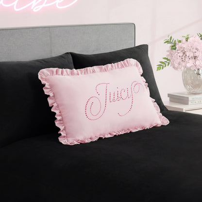 A pink Ruffle Decorative Pillow with rhinestone accents sits on a black bedspread, paired with a gray headboard and a vase of pink flowers on the bedside table in the background.