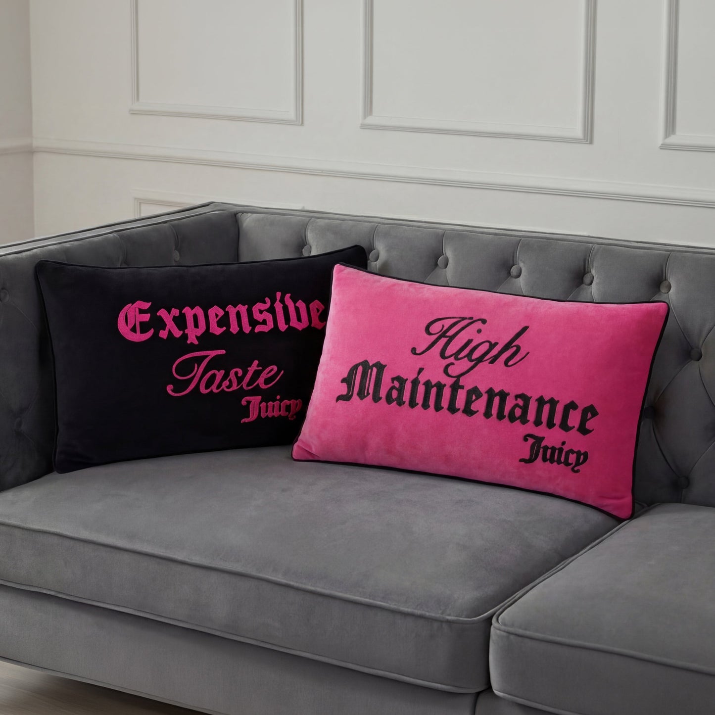 Two Reversible Decorative Pillows by Juicy Couture accent a gray tufted sofa: one black with pink “Expensive Taste Juicy,” the other pink with black “High Maintenance Juicy.” Both pop against a white paneled wall.