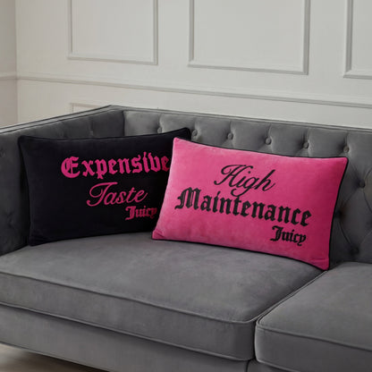 Two Reversible Decorative Pillows by Juicy Couture accent a gray tufted sofa: one black with pink “Expensive Taste Juicy,” the other pink with black “High Maintenance Juicy.” Both pop against a white paneled wall.