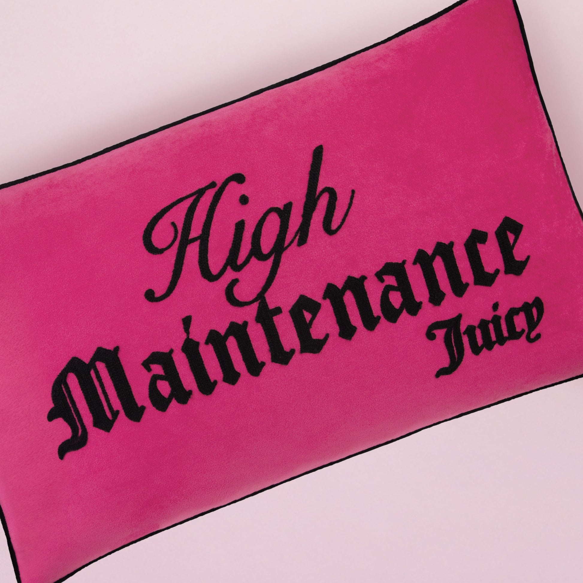 The reversible decorative pillow from Juicy Couture is pink and rectangular with black piping, featuring bold black "High Maintenance Juicy" text in a decorative font. It rests on a light pink surface.