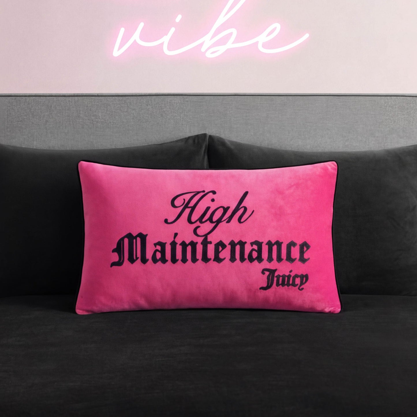 A bright pink Reversible Decorative Pillow with "High Maintenance Juicy" in black gothic font rests on a dark gray couch, with a glowing neon pink vibe sign on a light pink wall. This stylish accent adds playful energy to any space.