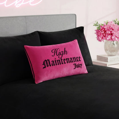 A vibrant pink Reversible Decorative Pillow with "High Maintenance Juicy" in black letters rests on a black bedspread beside a gray headboard, while pink flowers and books on the nightstand complete this chic setup.