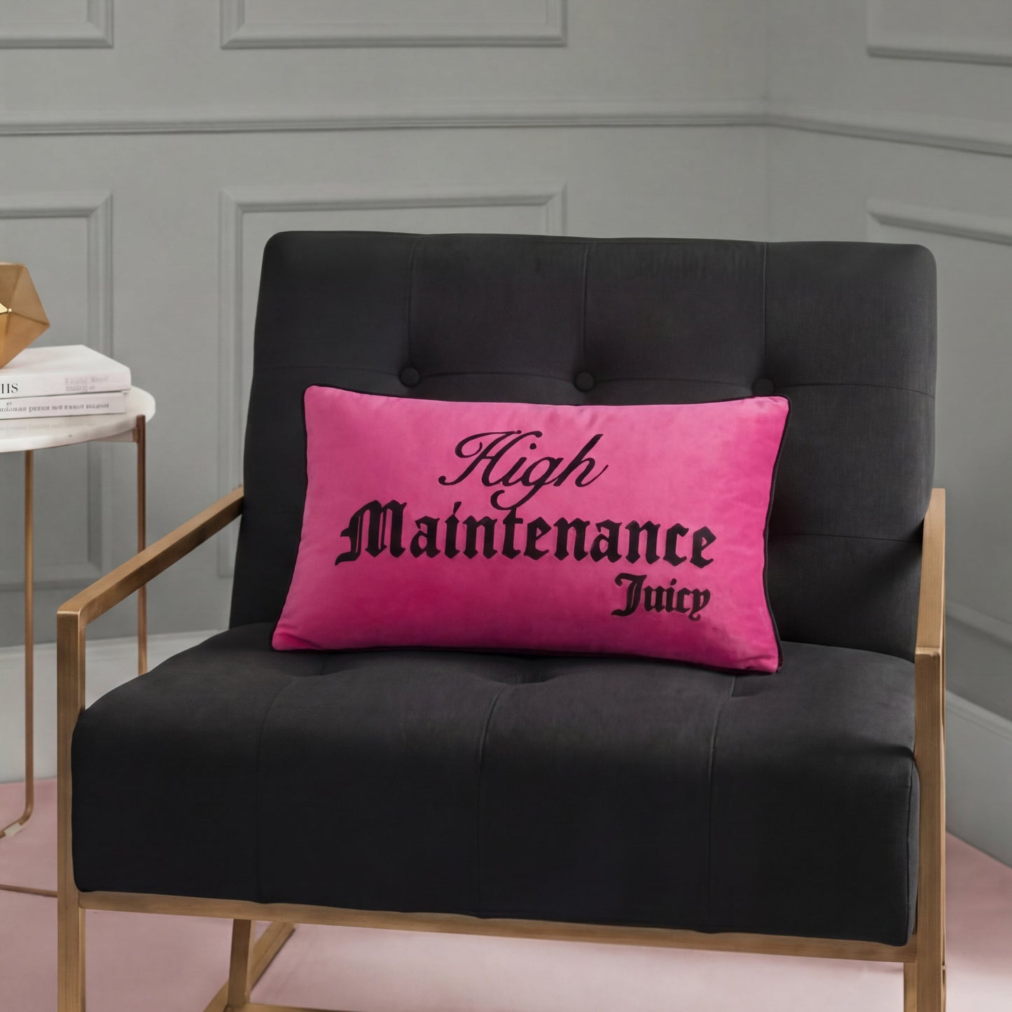 A black armchair with gold legs features a Reversible Decorative Pillow displaying “High Maintenance Juicy” in black script, set against a gray paneled wall and pink floor.