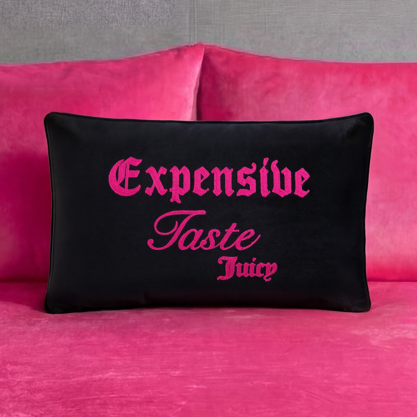 A black Juicy Couture Reversible Decorative Pillow with pink “Expensive Taste Juicy” text is placed on a bright pink velvet couch with matching cushions against a gray wall.