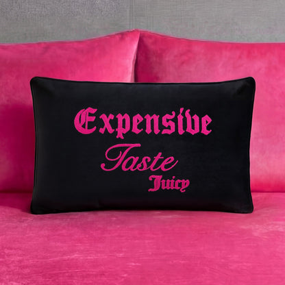 A black Juicy Couture Reversible Decorative Pillow with pink “Expensive Taste Juicy” text is placed on a bright pink velvet couch with matching cushions against a gray wall.