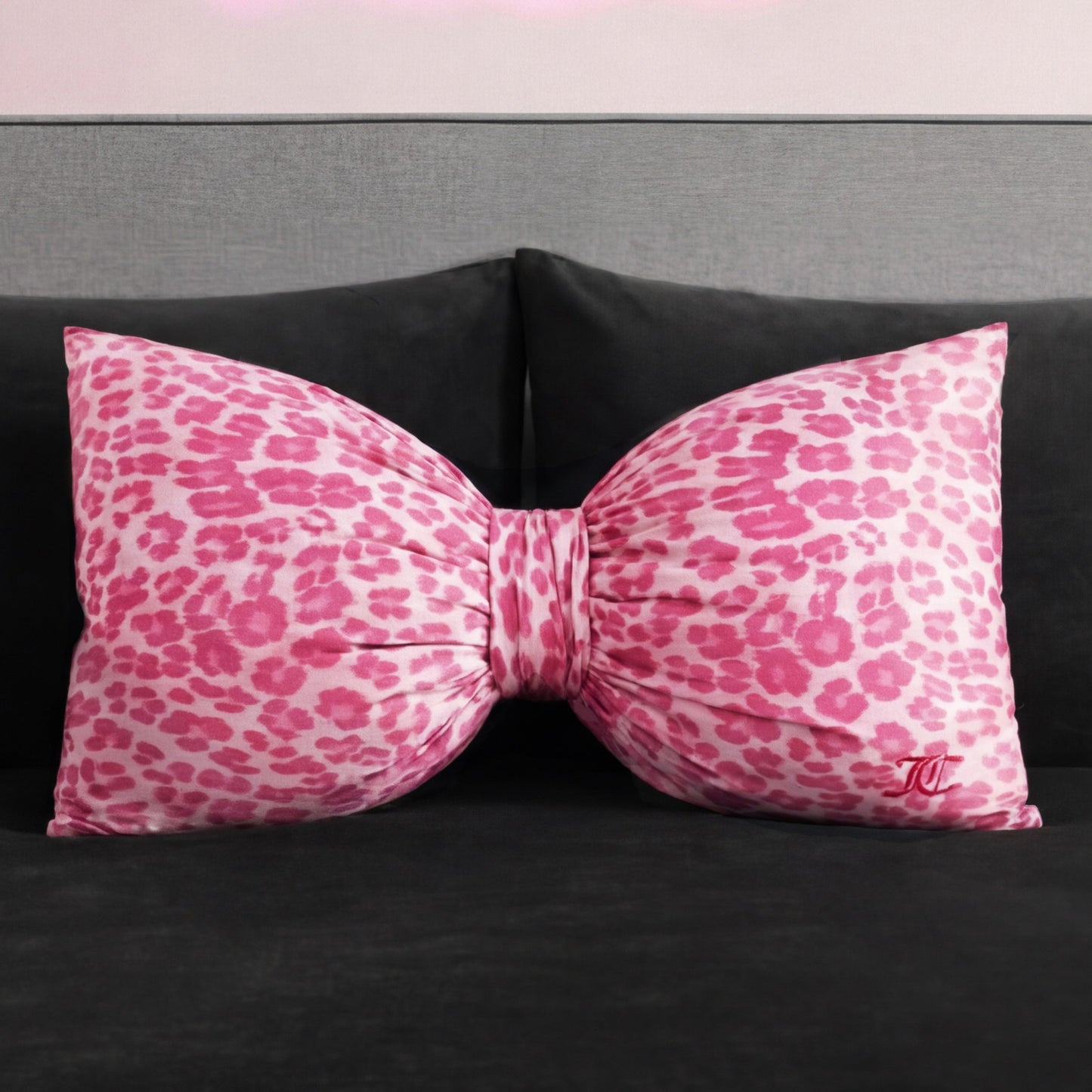 A Bow Decorative Pillow from Juicy Couture, featuring a pink and white leopard print and a small embroidered monogram, rests on a dark gray sofa or bed.