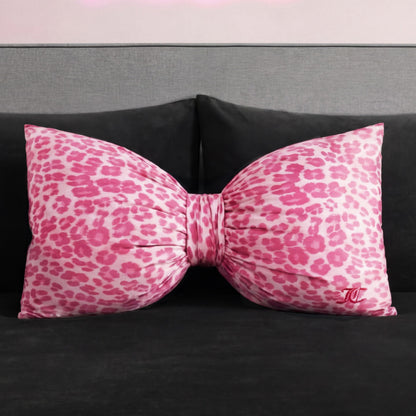 A Bow Decorative Pillow from Juicy Couture, featuring a pink and white leopard print and a small embroidered monogram, rests on a dark gray sofa or bed.