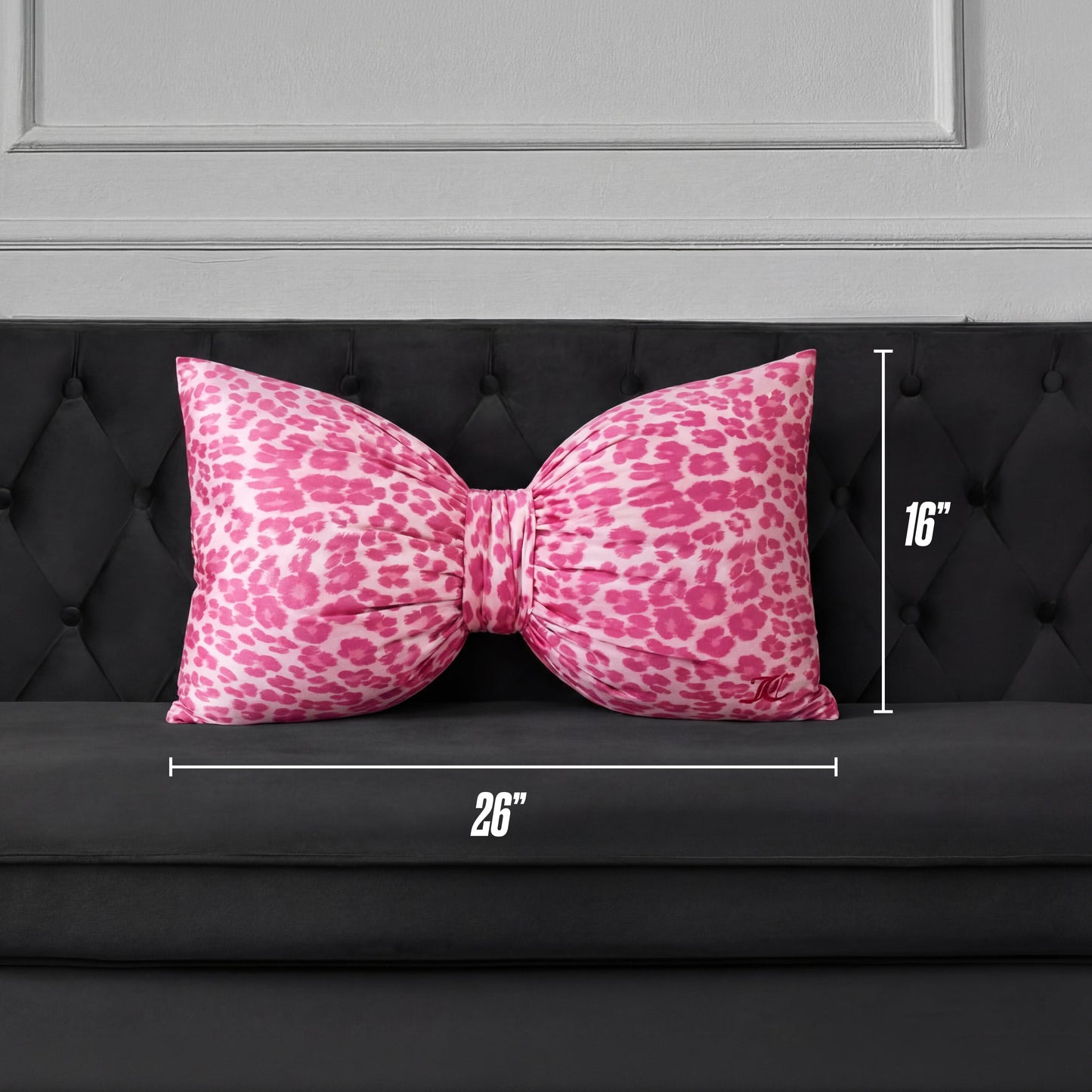 A Bow Decorative Pillow in pink cheetah print, sized 26" x 16", is placed on a black tufted sofa against a gray paneled wall.