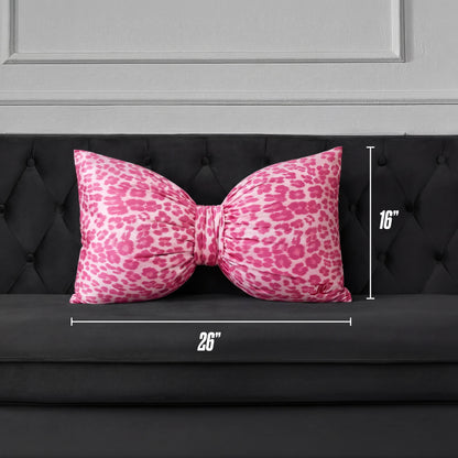 A Bow Decorative Pillow in pink cheetah print, sized 26" x 16", is placed on a black tufted sofa against a gray paneled wall.
