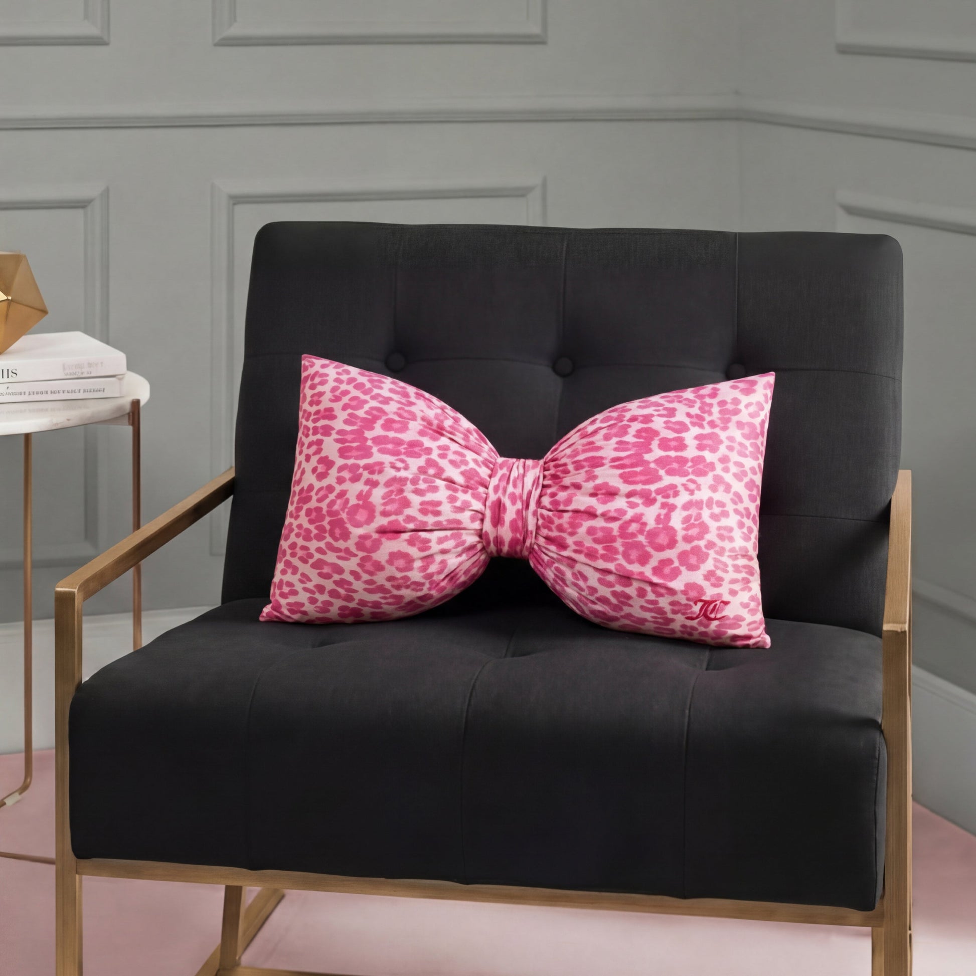A black armchair with gold legs holds a large Bow Decorative Pillow in cheetah print. Next to it is a small side table topped with books and a gold geometric decor piece.