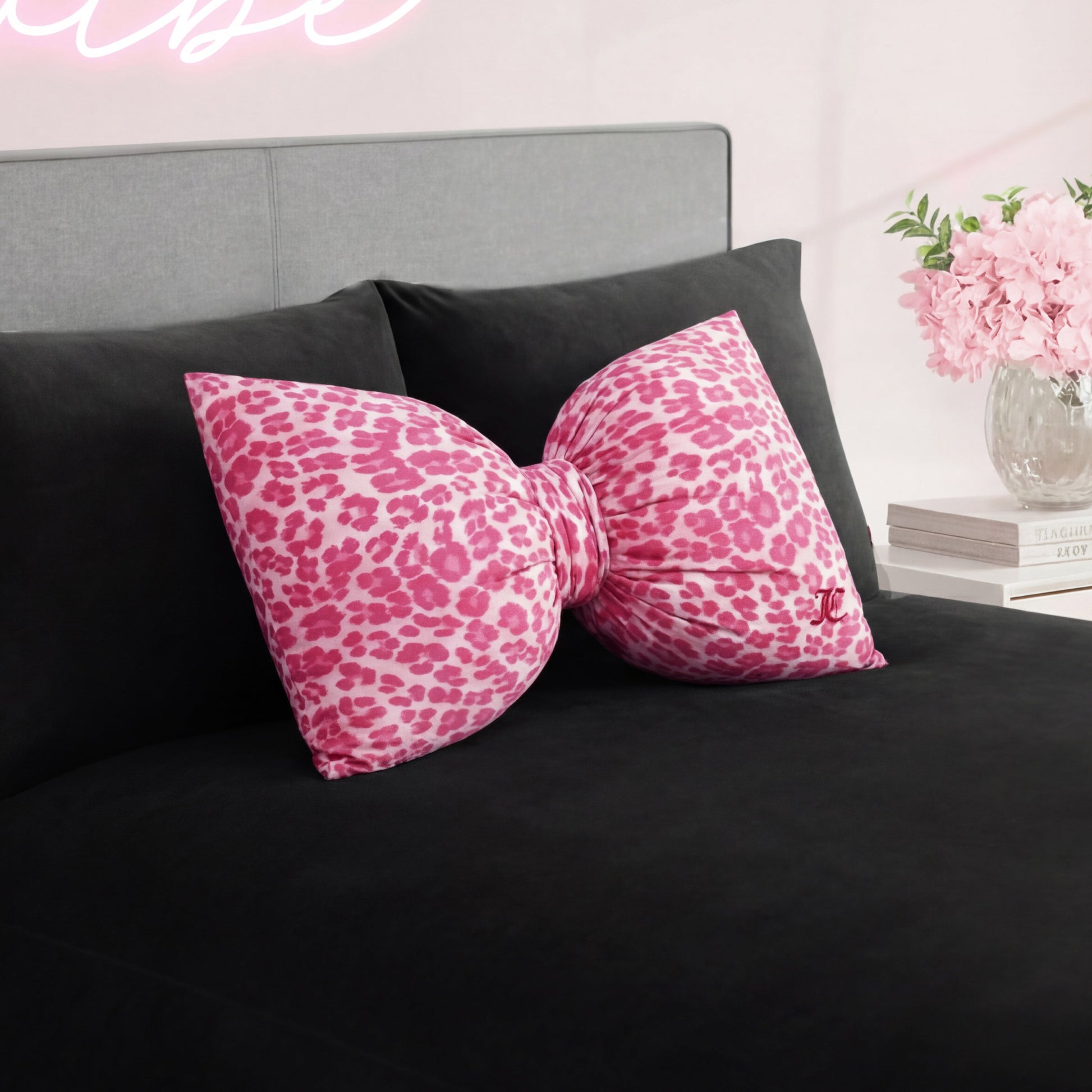 The Bow Decorative Pillow, featuring a large pink cheetah print design, adds a stylish touch to the black bedspread in a chic bedroom with light walls and a vase of pink flowers on the bedside table.