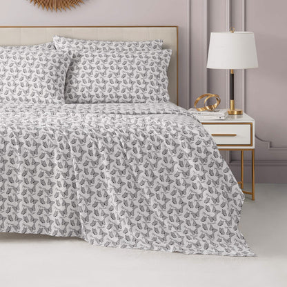 A neatly made bed with a Microfiber Sheet Set With Extra Pillowcases in white and gray leaf pattern, two pillows, and a matching comforter. Next to the bed sits a modern white nightstand with gold accents, lamp, books, and décor.