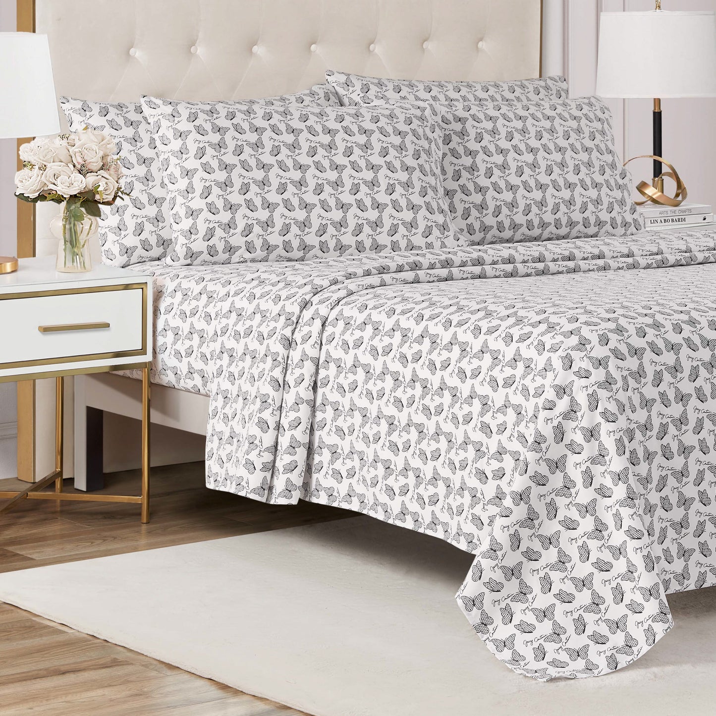 A neatly made bed features the Microfiber Sheet Set With Extra Pillowcases in white and gray floral print, next to a white nightstand with gold accents, a vase of flowers, and a modern lamp in a bright, elegant bedroom.