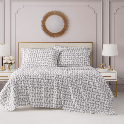 A stylish bedroom features a neatly made bed with the Microfiber Sheet Set With Extra Pillowcases in white and gray, centered among light gray paneled walls, a round sunburst mirror, white nightstands, lamps, and a vase of flowers.