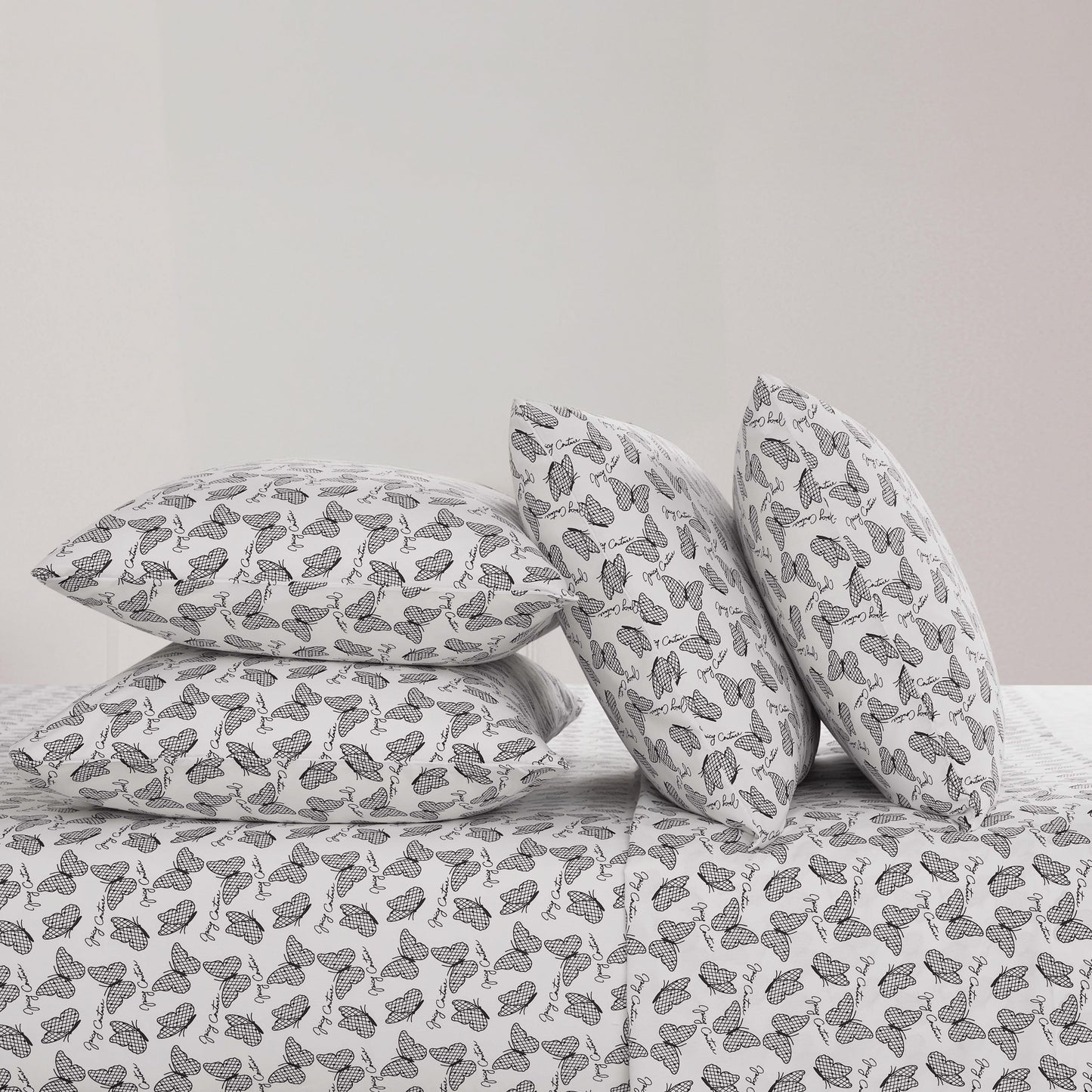 Four black and white butterfly patterned pillows from the Microfiber Sheet Set With Extra Pillowcases are stacked on a luxury bedspread—two upright and two flat—against a plain, light-colored wall.