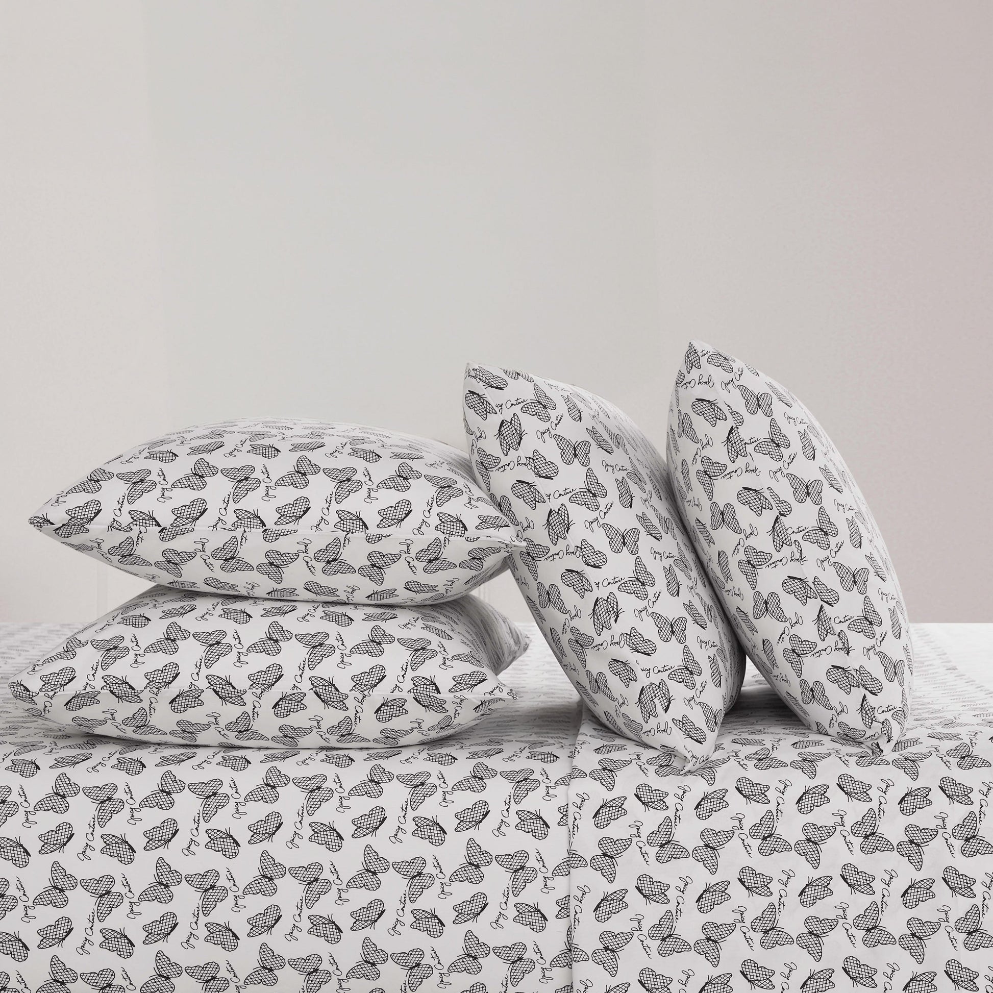 Four black and white butterfly patterned pillows from the Microfiber Sheet Set With Extra Pillowcases are stacked on a luxury bedspread—two upright and two flat—against a plain, light-colored wall.