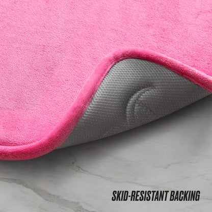 A pink Quilted Memory Foam Bath Mat 2-Piece Set with one corner lifted shows its gray, textured skid-resistant backing on a light floor. "SKID-RESISTANT BACKING" is visible in the bottom right corner.