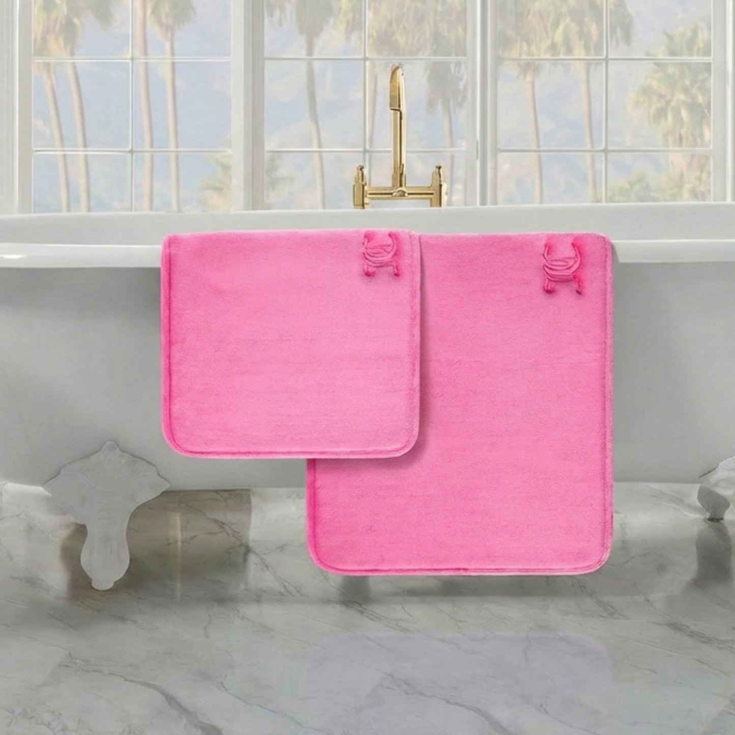 Two pink Quilted Memory Foam Bath Mats hang over a white clawfoot tub with a gold faucet. Large windows reveal palm trees outside, while a marble floor enhances the bathroom’s luxurious feel.