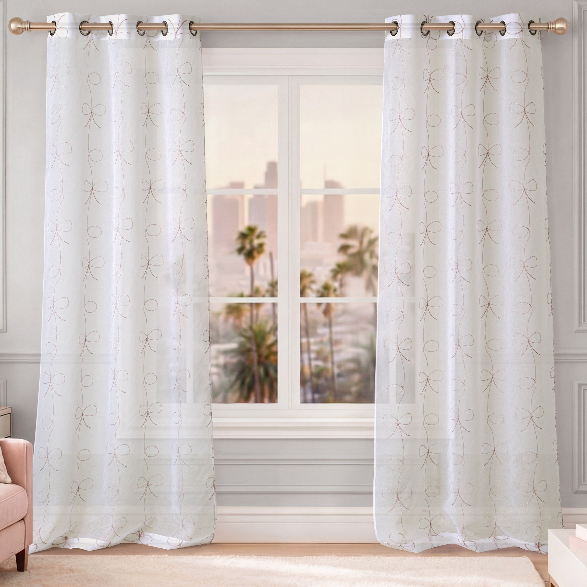 Two sheer white Vertical Bow Ribbon Sheer Window Curtains with a subtle loop pattern hang from a gold curtain rod in front of a window. Palm trees and a city skyline are visible outside. A blush pink chair is partially visible on the left.