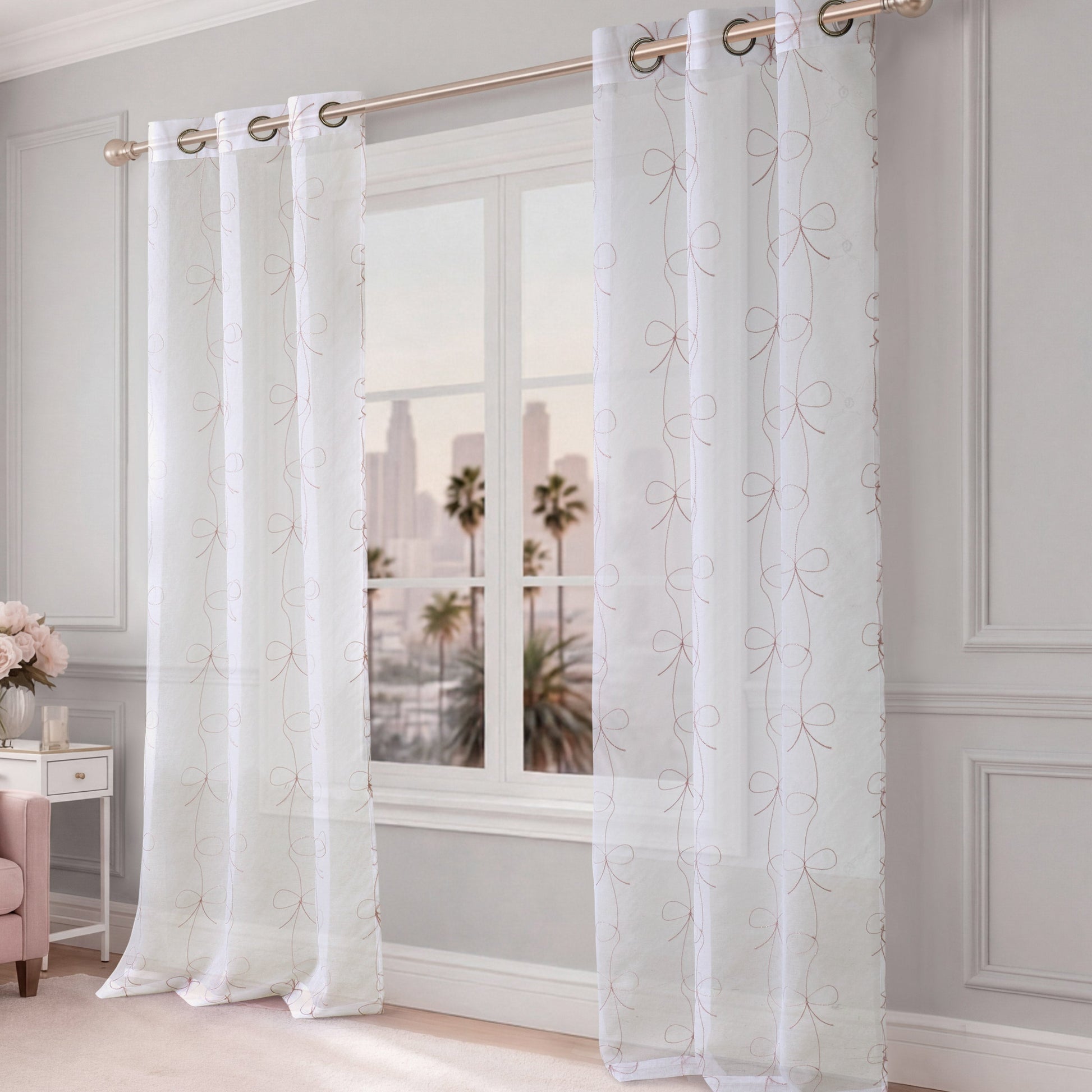 Vertical Bow Ribbon Sheer Window Curtains hang from a gold curtain rod in front of a window that overlooks a cityscape with palm trees. The room features light gray walls and elegant decor, creating a chic atmosphere reminiscent of Juicy Couture’s signature style.