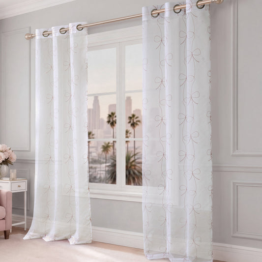 Vertical Bow Ribbon Sheer Window Curtains hang from a gold curtain rod in front of a window that overlooks a cityscape with palm trees. The room features light gray walls and elegant decor, creating a chic atmosphere reminiscent of Juicy Couture’s signature style.