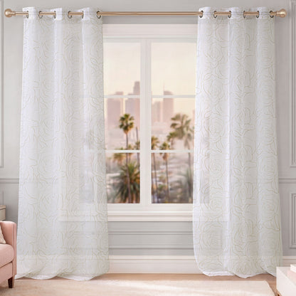 A set of Rose Swirl Sheer Window Curtains with embroidered floral patterns hangs on a gold curtain rod in front of the window. Palm trees and city buildings can be seen outside, while light walls and a pink chair in the corner enhance the room’s airy, elegant ambiance.