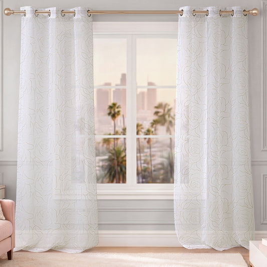 A set of Rose Swirl Sheer Window Curtains with embroidered floral patterns hangs on a gold curtain rod in front of the window. Palm trees and city buildings can be seen outside, while light walls and a pink chair in the corner enhance the room’s airy, elegant ambiance.