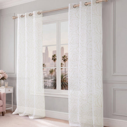 Rose Swirl Sheer Window Curtains, adorned with delicate white embroidered floral and subtle rose patterns, hang from a gold rod in front of a large window. Through the glass, palm trees and buildings can be seen. The room features light gray walls and elegant decor.