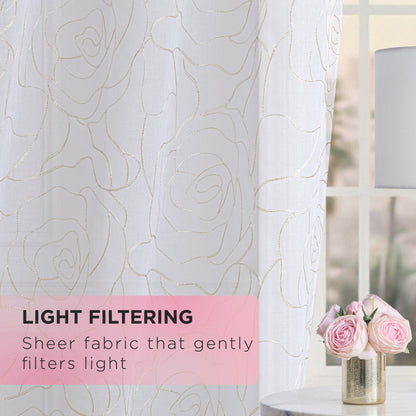 Rose Swirl Sheer Window Curtains, adorned with a delicate rose pattern, gently filter sunlight by the window for an effortlessly elegant look. On a nearby table sit a vase of pink roses and a white lamp. These embroidered floral curtains feature a convenient grommet header for easy hanging. Text reads: “Light Filtering. Sheer fabric that gently filters light.”.