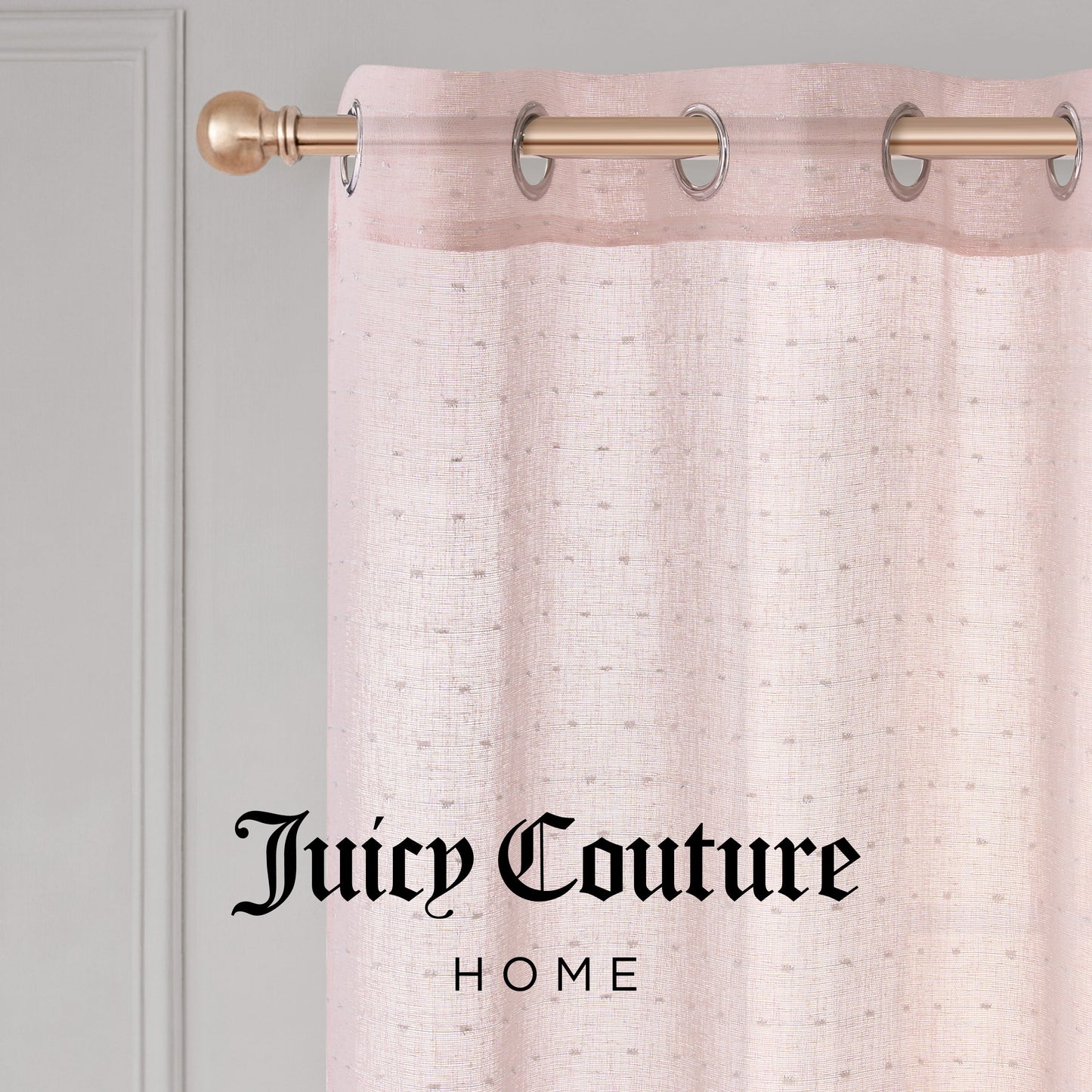 Close-up of the Aurora Sheer Window Curtain by Juicy Couture, featuring a pale pink sheer fabric with a grommet header on a gold rod. "Juicy Couture Home" is printed in black text on the curtain.