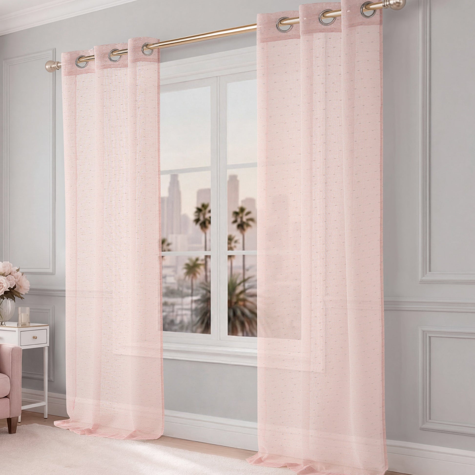 The Aurora Sheer Window Curtain in pink with a grommet header hangs from a gold rod, allowing natural light to fill the room. A cityscape with palm trees appears beyond the window, complementing the light gray walls and elegant decor.
