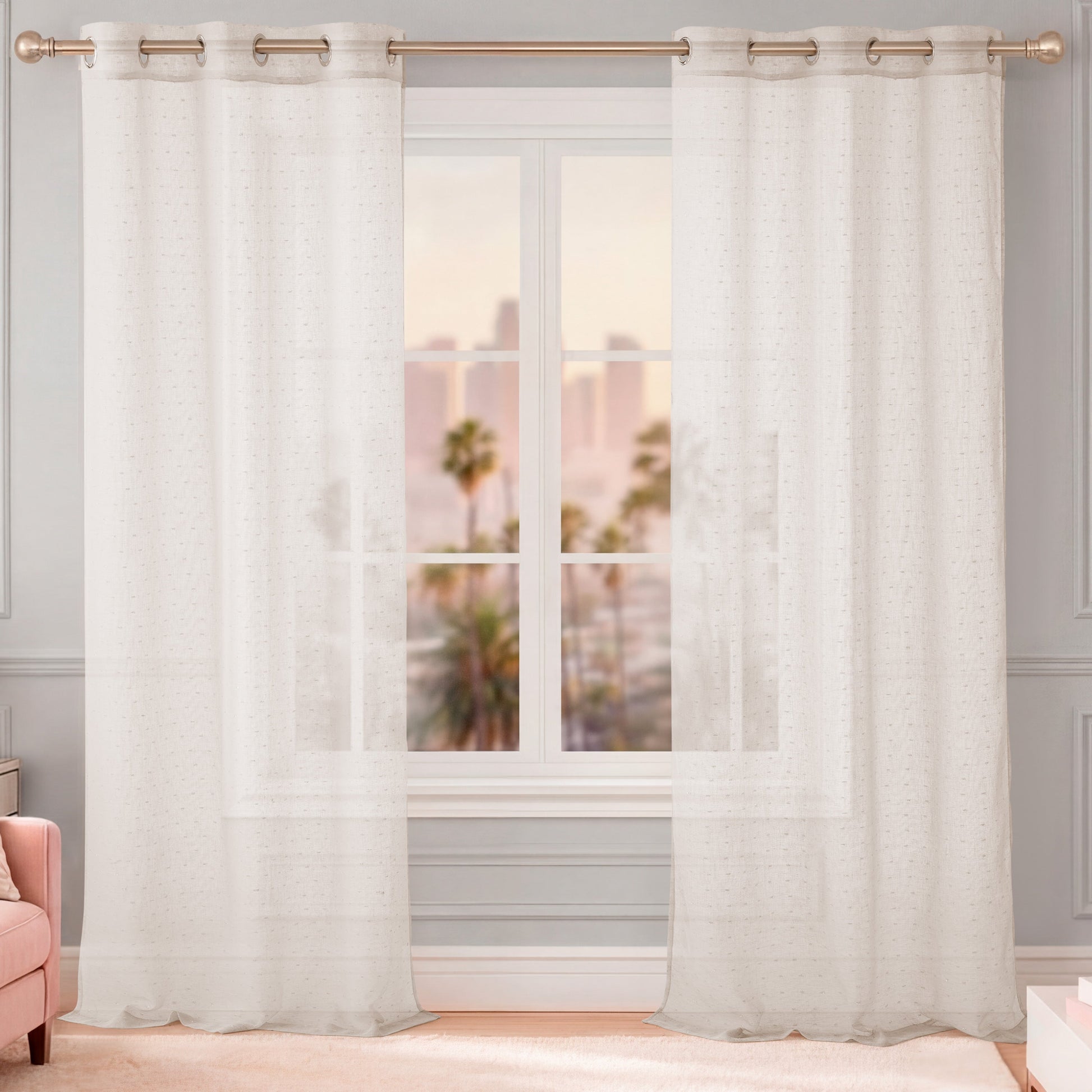 The Aurora Sheer Window Curtain with a grommet header hangs on a rod, allowing sunlight to filter through. Blurry palm trees and buildings are visible outside, and a pink chair is partially seen on the left.