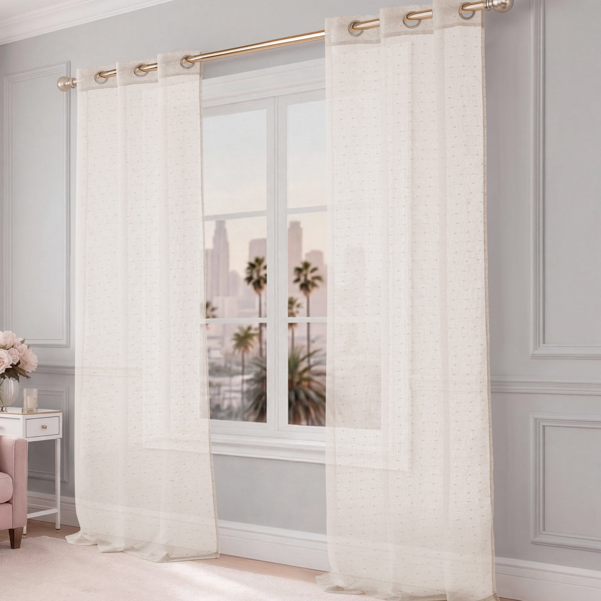 The Aurora Sheer Window Curtain hangs on a gold rod above the window, revealing views of palm trees and city buildings. The room has light gray walls, elegant decor, a pink chair, and flowers.