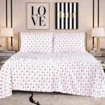 A bed dressed in the Microfiber Sheet Set With Extra Pillowcases, featuring white sheets with a red and black rose pattern, is flanked by two nightstands topped with lamps. Above the bed hang two wall art pieces: one that reads "LOVE" and another that says "Juicy" on black and white stripes.