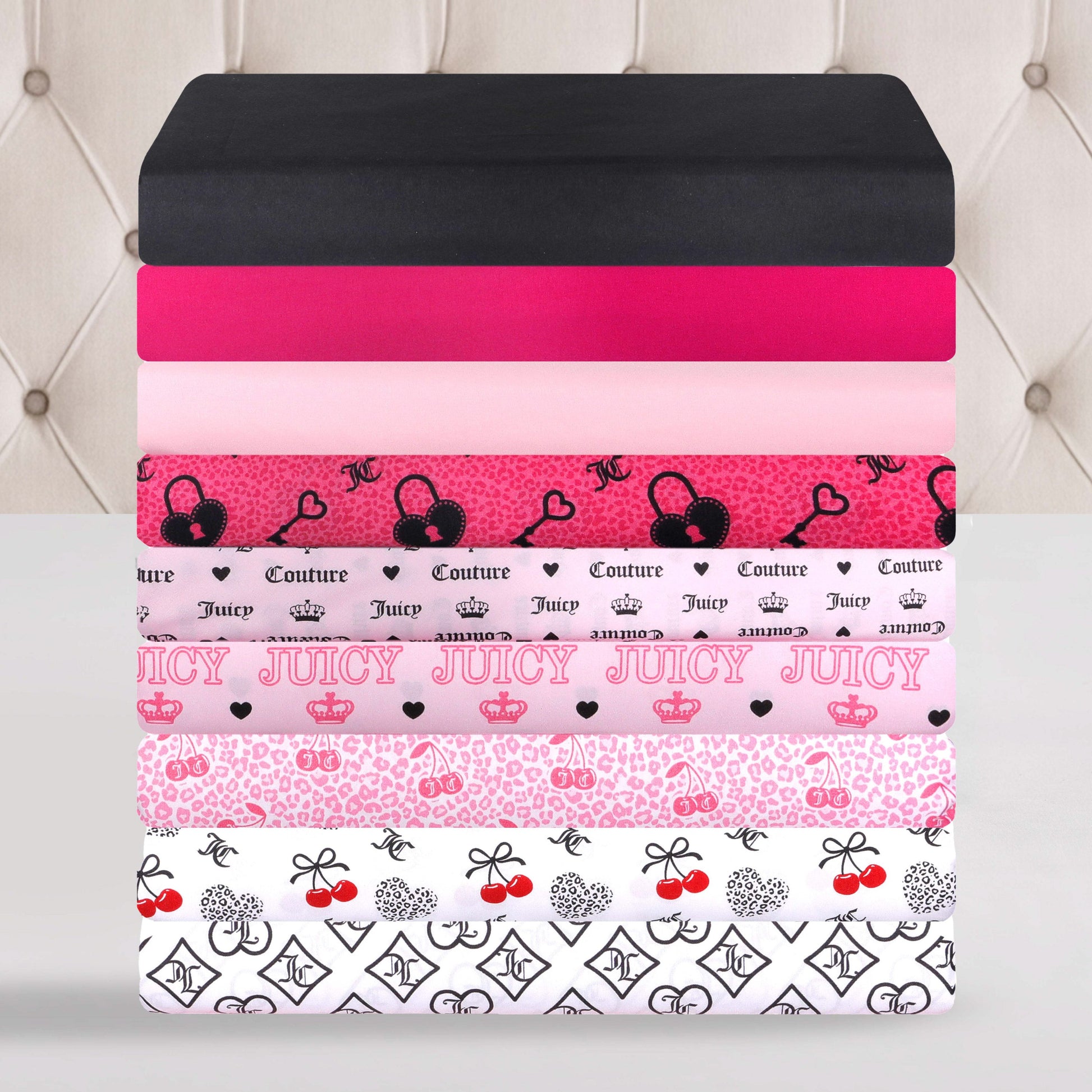 A stack of folded sheets in shades of black, pink, and white—adorned with lively Juicy Couture-inspired patterns like hearts, cherries, crowns, and bows—adds a vibrant accent against a tufted beige headboard. The Microfiber Sheet Set With Extra Pillowcases combines playful style with luxurious comfort for your space.