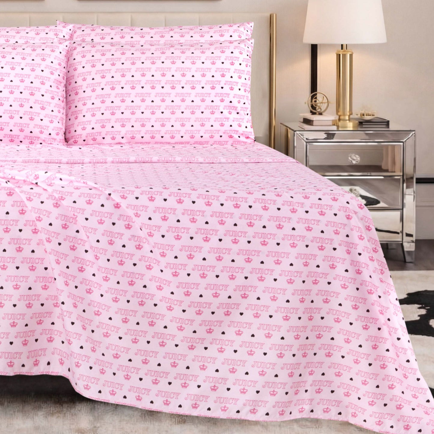 The pink bedspread and pillows showcase repeating JUICY text with heart and lips patterns, perfectly capturing the playful essence of Juicy Couture bedding. The bed is complemented by the Microfiber Sheet Set With Extra Pillowcases, adding an ultra-soft touch. A mirrored nightstand with a lamp and décor completes the modern bedroom setting.