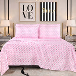 A bed with pink heart-patterned Juicy Couture bedding and a Microfiber Sheet Set With Extra Pillowcases sits between two mirrored nightstands with lamps. Behind the bed are two framed wall art pieces, one reading LOVE and the other Juicy on a striped background.