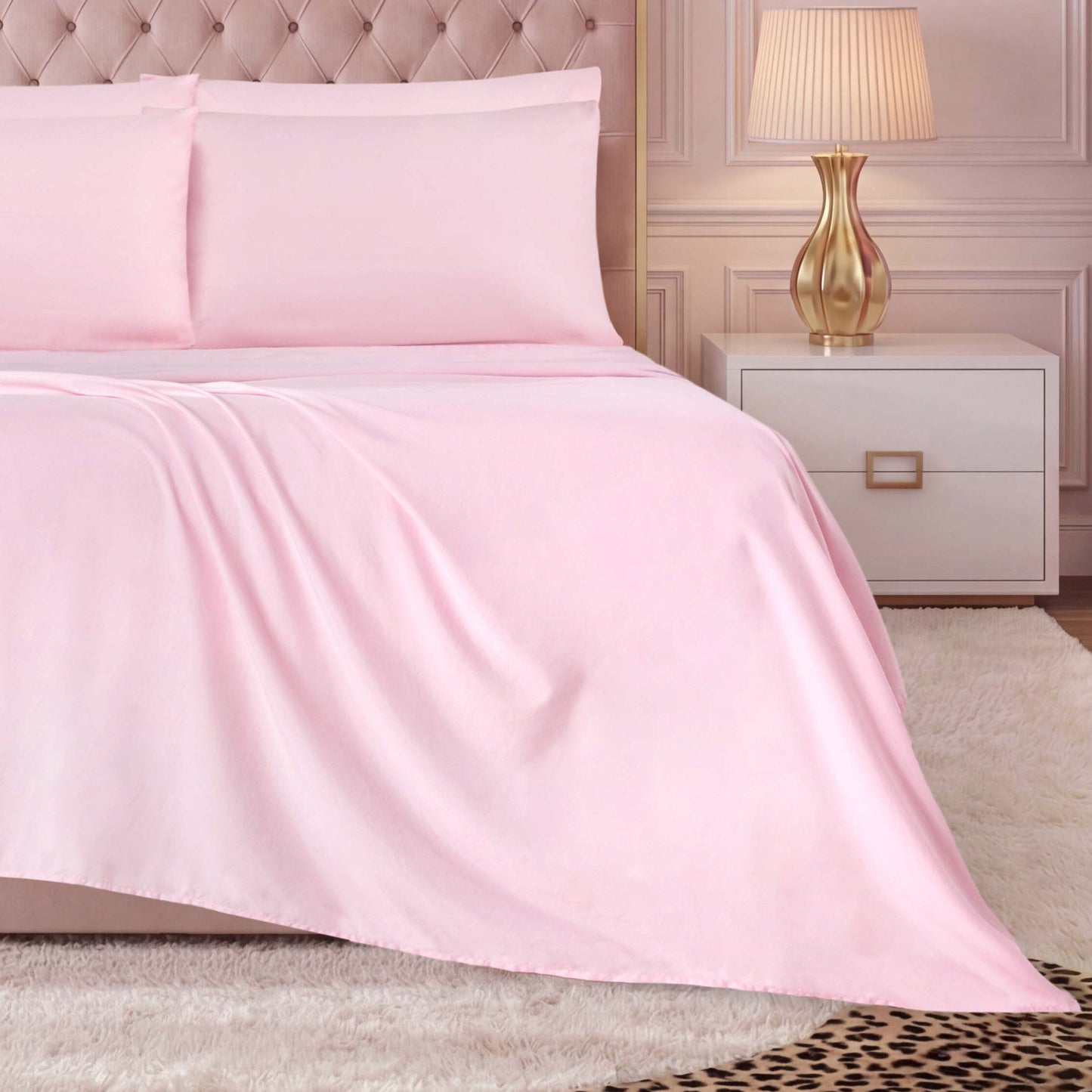 A neatly made bed with light pink luxury bedding, including a Microfiber Sheet Set With Extra Pillowcases and matching pillows, a white nightstand with a gold lamp, and a soft white rug on the floor next to a leopard-print rug.
