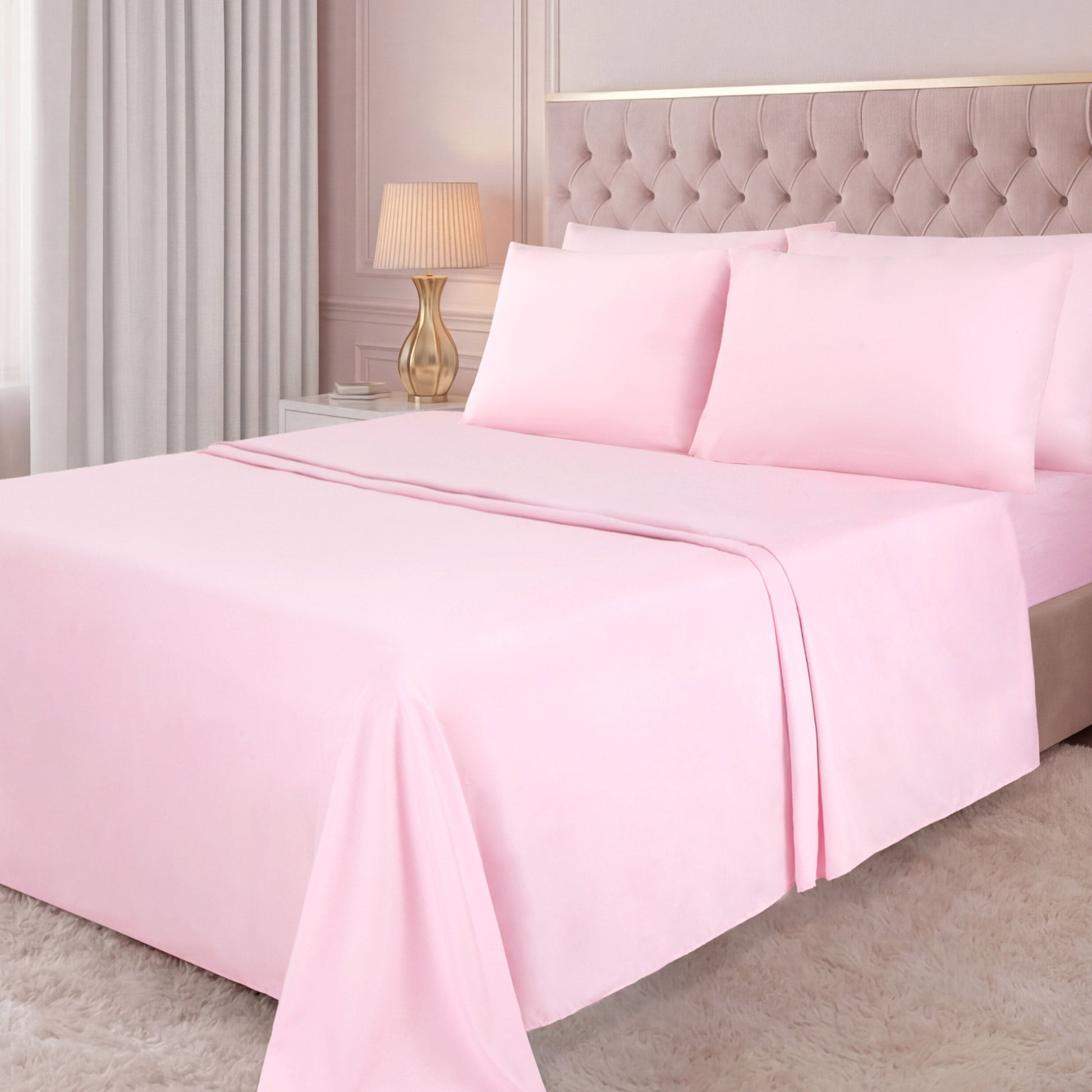 A neatly made bed with a light pink Microfiber Sheet Set With Extra Pillowcases, a tufted beige headboard, a white nightstand with a gold lamp, light curtains, and a white shaggy rug in a softly lit bedroom create an inviting atmosphere of luxury bedding.