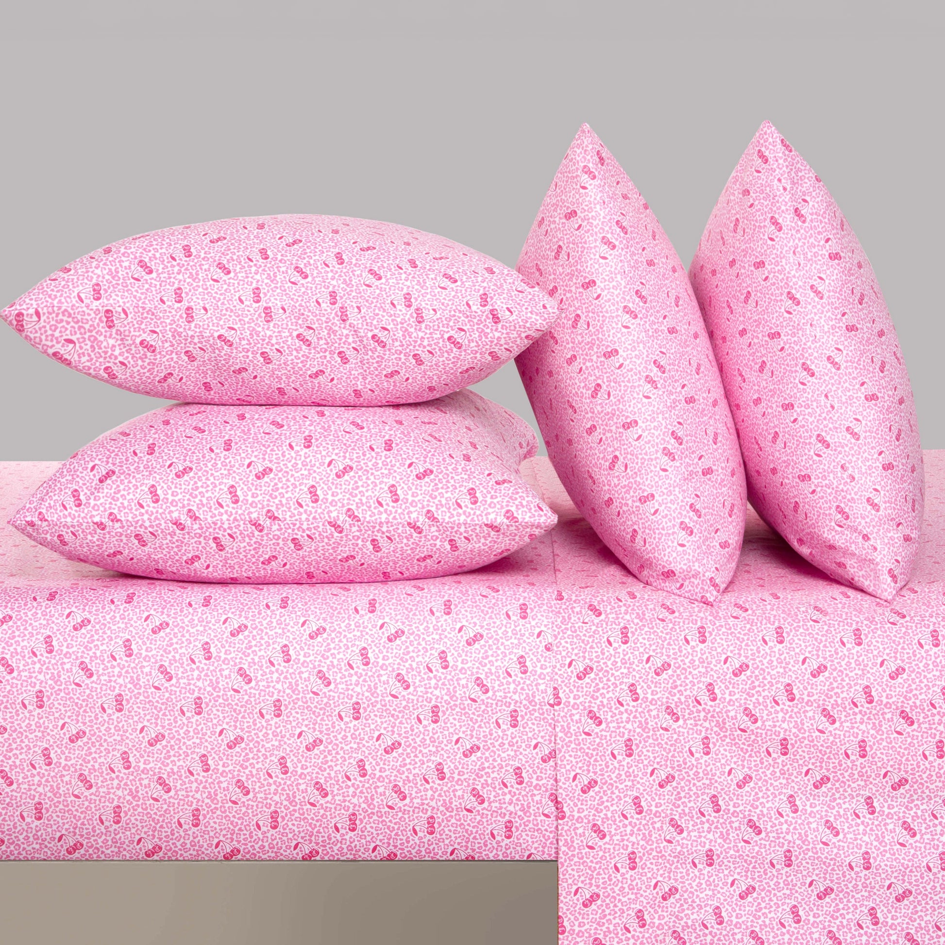 Four pink pillows with a small, repeating pattern are stacked and propped up on a matching Microfiber Sheet Set With Extra Pillowcases, all set against a plain light gray background for a touch of luxury bedding perfection.